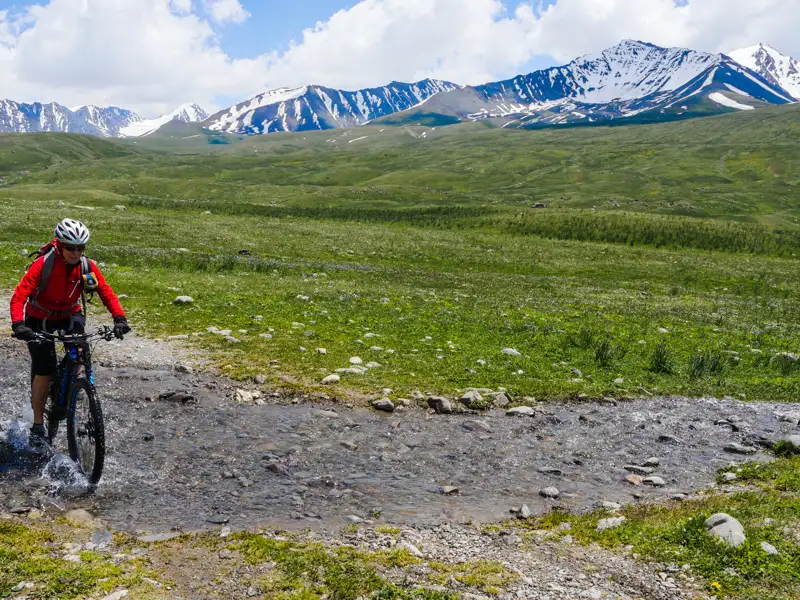 Mountainbiker durchquert einen Bach im Hochgebirge vor der Kulisse schneebedeckter Gipfel.