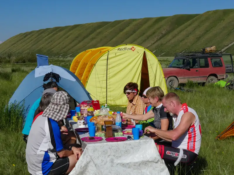 Reiseteilnehmer beim Mittagessen an einem Tisch vor Zelten. Ein Geländewagen steht im Hintergrund.