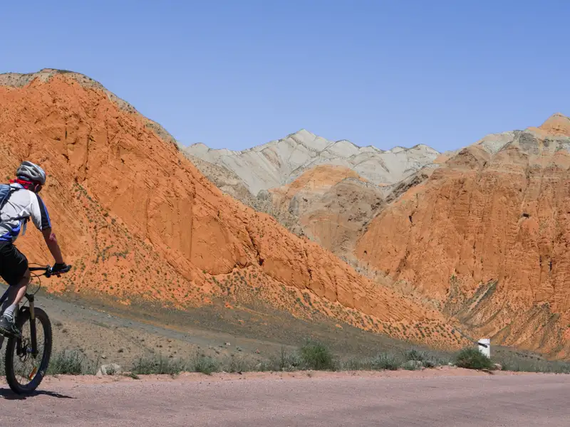 Mountainbiker auf einer Straße mit roten und beigefarbenen Bergen im Hintergrund.