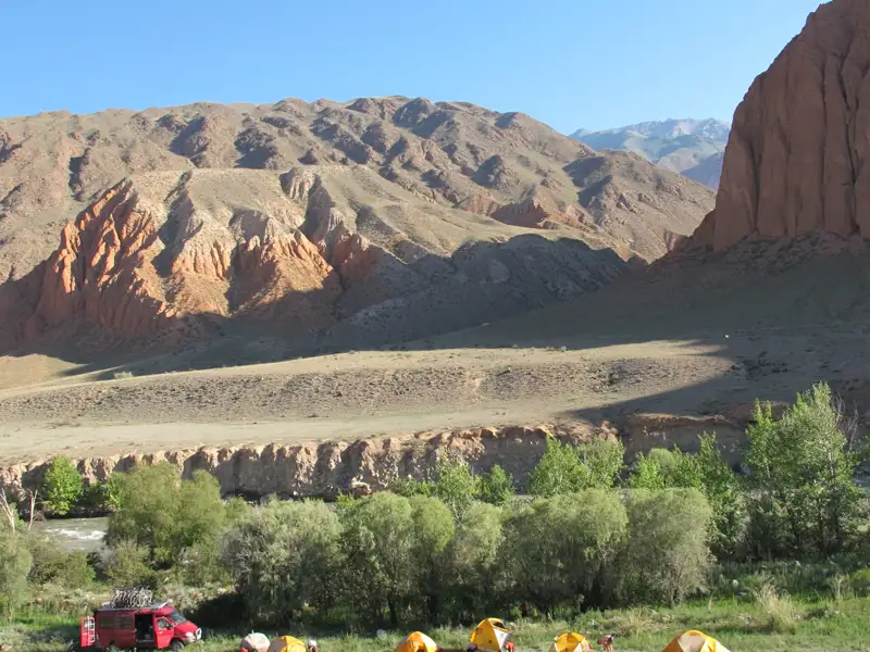 Campingplatz in einer Berglandschaft. Zelte stehen am Ufer eines Flusses, ein roter Van ist in der Nähe geparkt. Im Hintergrund sind rötliche Felsformationen und Berge zu sehen.