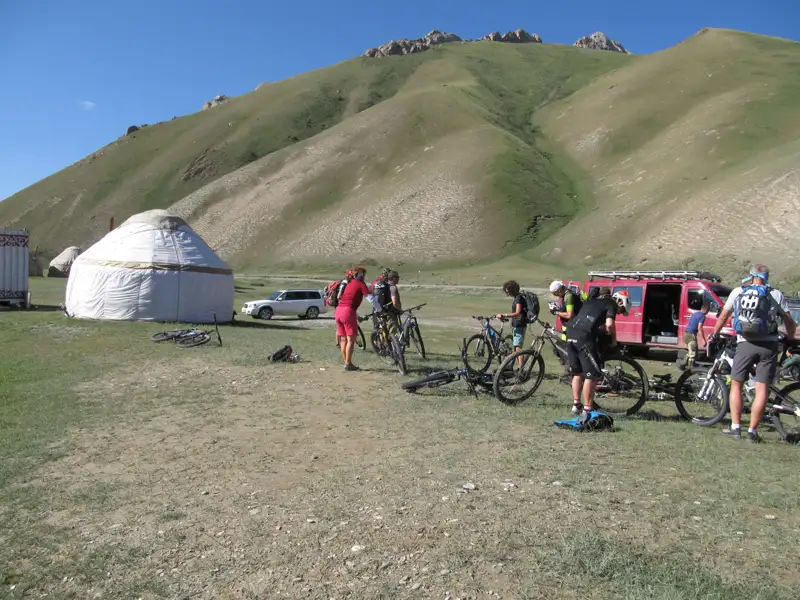 Mountainbiker legen eine Pause während ihrer Tour ein. Sie stehen mit ihren Fahrrädern vor einer Jurte, die vermutlich als Unterkunft dient. Ein Begleitfahrzeug ist ebenfalls zu sehen.