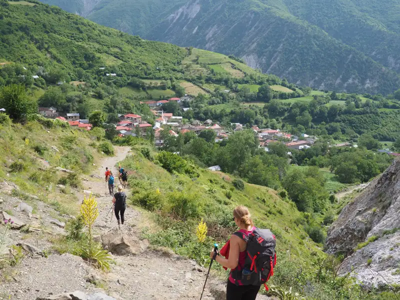 Wanderer mit Rucksäcken und Wanderstöcken auf einem Bergpfad. Im Hintergrund liegt ein Dorf in einem grünen Tal.