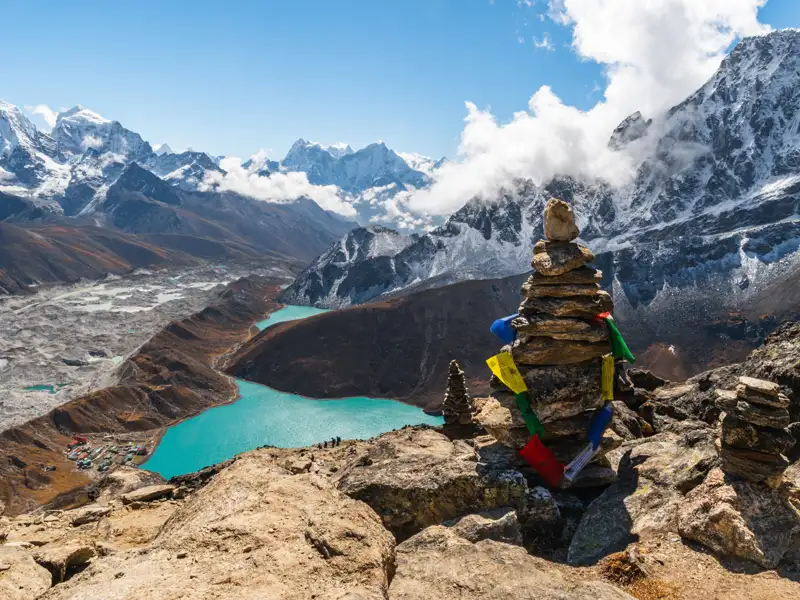 Türkisfarbene Gletscherseen im Himalaya mit Blick auf die schneebedeckten Gipfel und Gebetsfahnen an einem Steinhaufen.