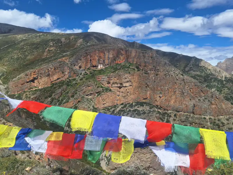 Gebetsfahnen im Vordergrund, dahinter eine Berglandschaft mit einem Gebäude, möglicherweise ein Kloster oder Tempel.