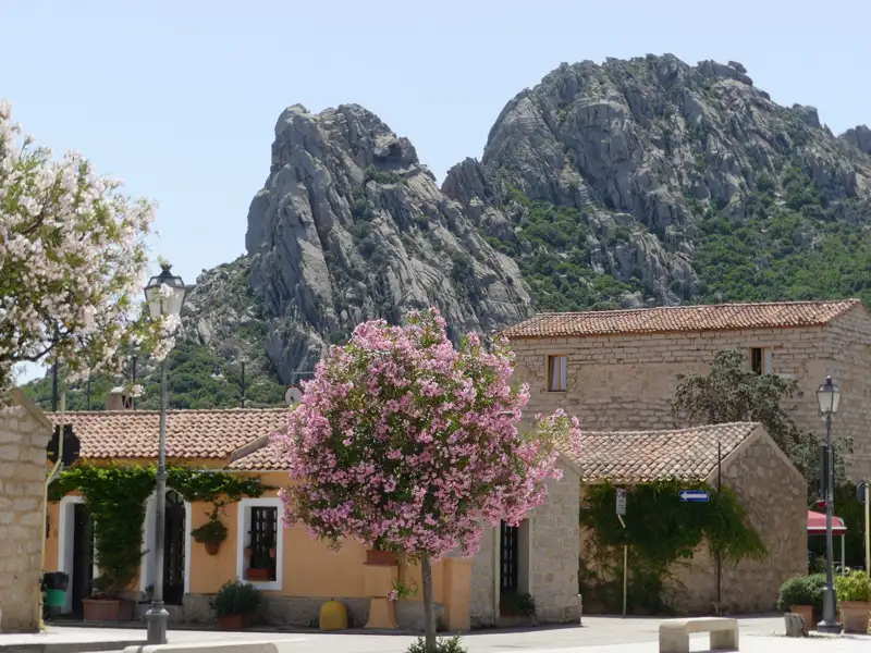 Szene eines Dorfplatzes mit traditionellen Häusern und einem großen Felsen im Hintergrund.