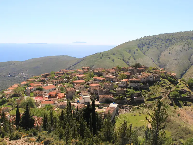 Panoramablick auf ein Bergdorf mit traditionellen Häusern, das sich an einen Hügel schmiegt. Im Hintergrund erstreckt sich das blaue Meer.