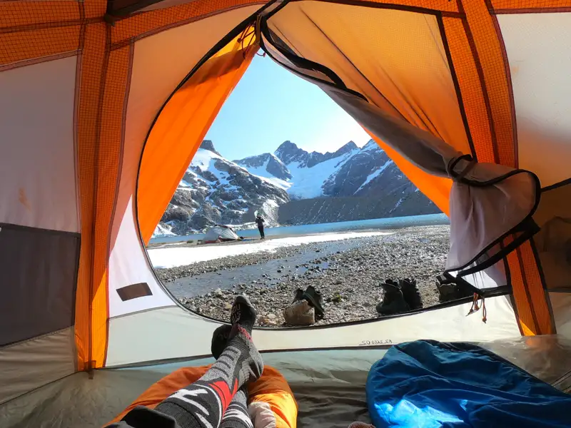 Blick aus einem Zelt auf eine Berglandschaft mit Schnee, einem See oder Fluss und einer Person, die im Zelt liegt.