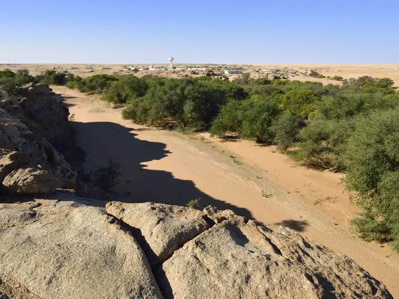 Trockenes Flussbett mit spärlicher Vegetation in einer ariden Landschaft. Eine Siedlung im Hintergrund zeigt die Anpassung an die trockenen Bedingungen.