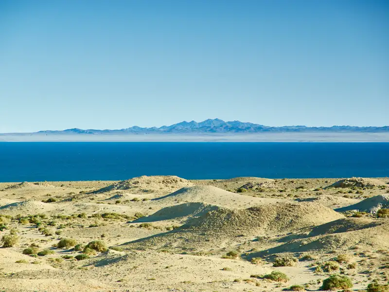 Landschaft mit sandigen Hügeln, blauem Wasser und Bergen im Hintergrund.