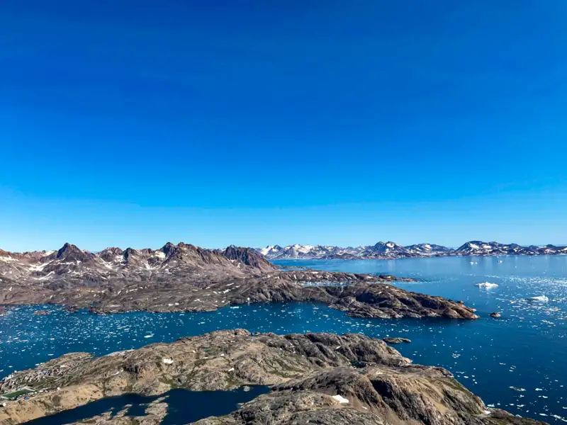 Felsige Küste Grönlands mit Eisbergen im Wasser und Bergen im Hintergrund.
