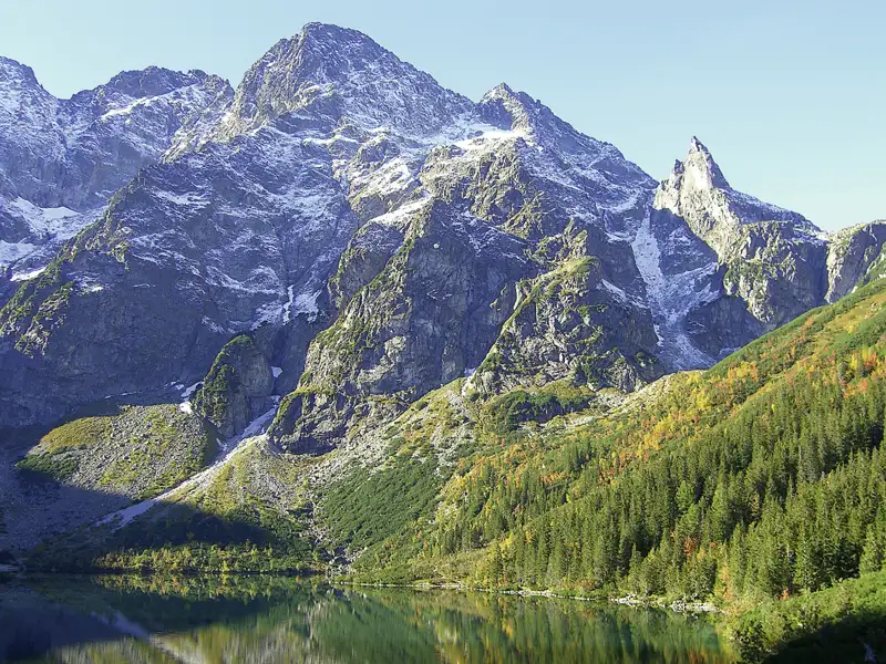 Blick auf einen Bergsee mit schneebedeckten Gipfeln und herbstlichem Wald.