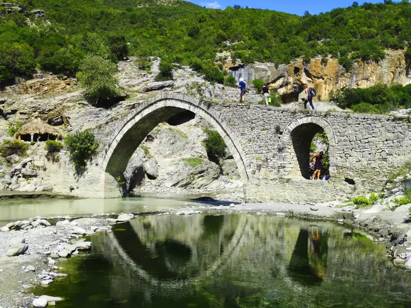Historische Steinbrücke mit Wanderern, Fluss und bewaldeter Landschaft im Hintergrund.