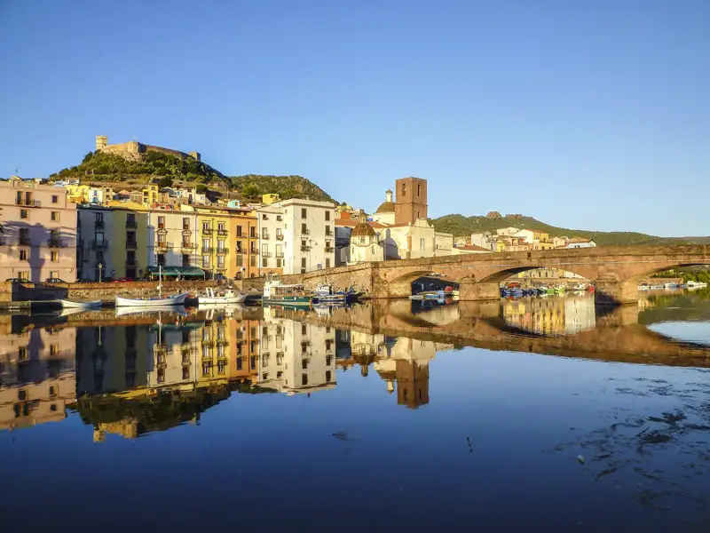 Bunte Häuser der Altstadt spiegeln sich im Fluss, im Hintergrund eine Brücke und eine Burg.