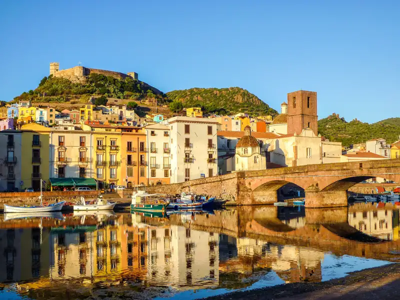 Farbenfrohe Häuserfassaden spiegeln sich im Wasser, eine Brücke führt über den Fluss, und im Hintergrund thront eine Burg auf einem Hügel.