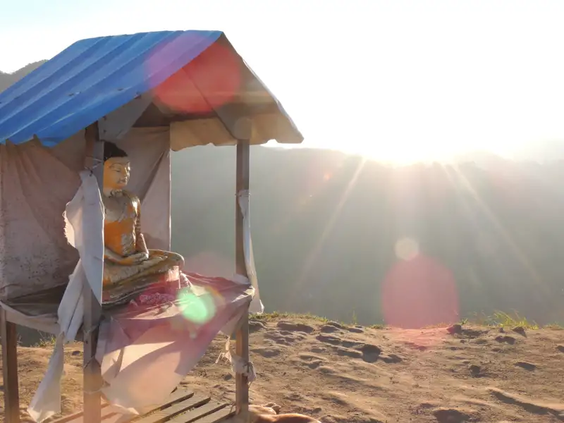 Eine Buddha-Statue unter einem Schutzdach mit Blick auf einen sonnigen Berghang.
