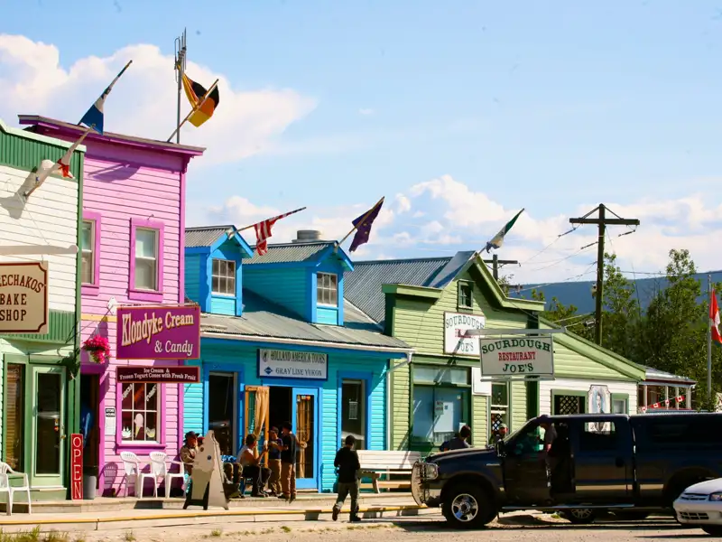 Bunt gestrichene Häuserfassaden von Geschäften und Restaurants in einer Stadt. Zu sehen sind unter anderem 'Klondyke Cream & Candy', 'Sourdough Joe's Restaurant' und 'Chechakos Bake Shop'. Ein schwarzer Pickup-Truck steht auf der Straße.
