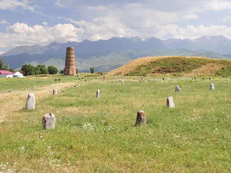 Burana-Turm in Kirgisistan mit Steinen und Hügeln im Vordergrund und Bergen im Hintergrund.