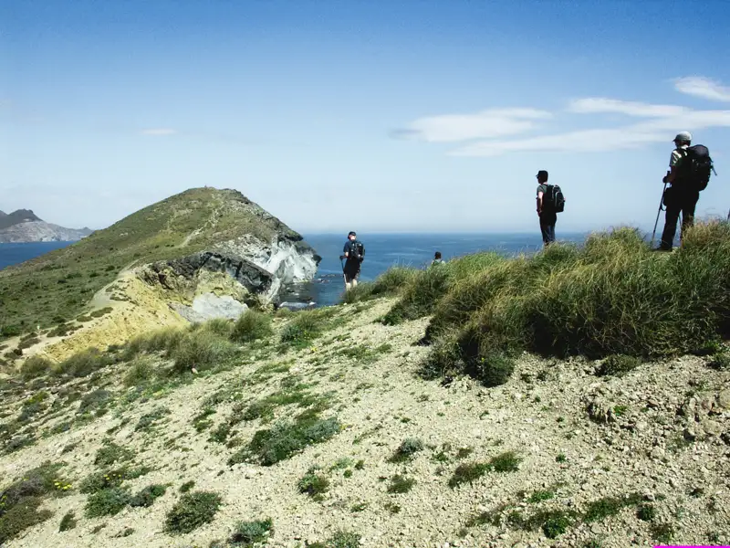 Wanderer mit Rucksäcken auf einem Küstenpfad, der Vegetation und felsiges Gelände zeigt, mit Blick auf das Meer und eine vorgelagerte Insel.