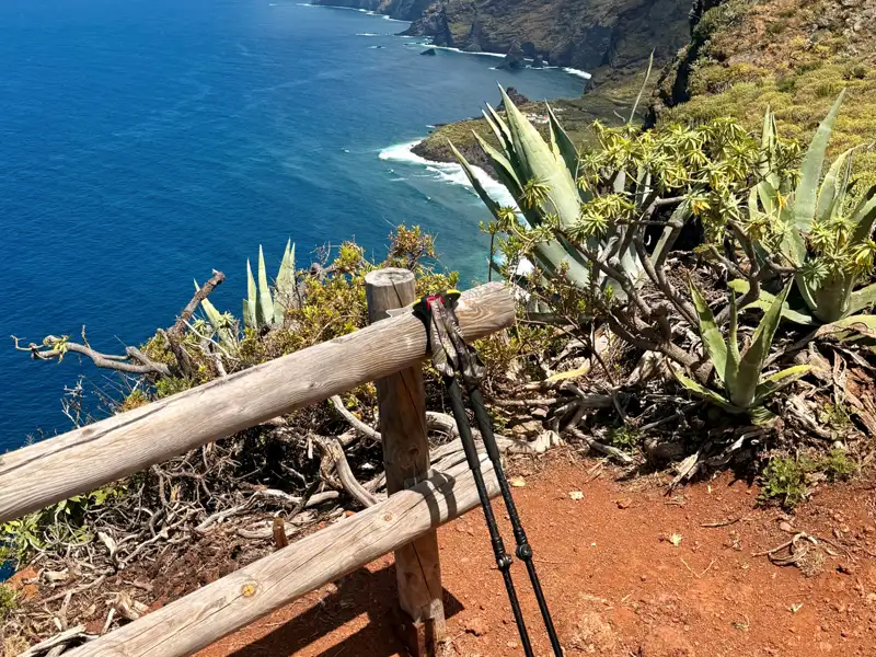 Wanderstöcke lehnen an einem hölzernen Geländer auf einem Küstenwanderweg. Im Hintergrund ist das blaue Meer und die felsige Küste zu sehen.