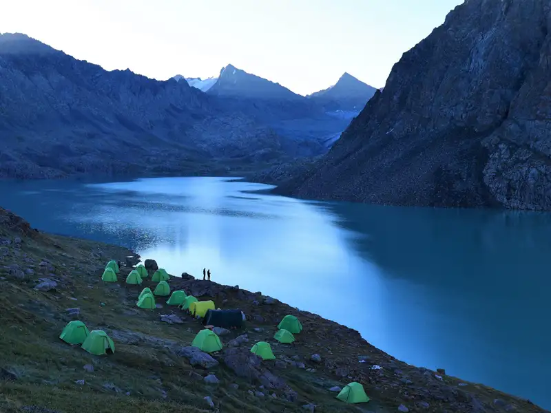 Campingplatz mit mehreren Zelten am Ufer eines türkisfarbenen Bergsees, umgeben von hohen Bergen.