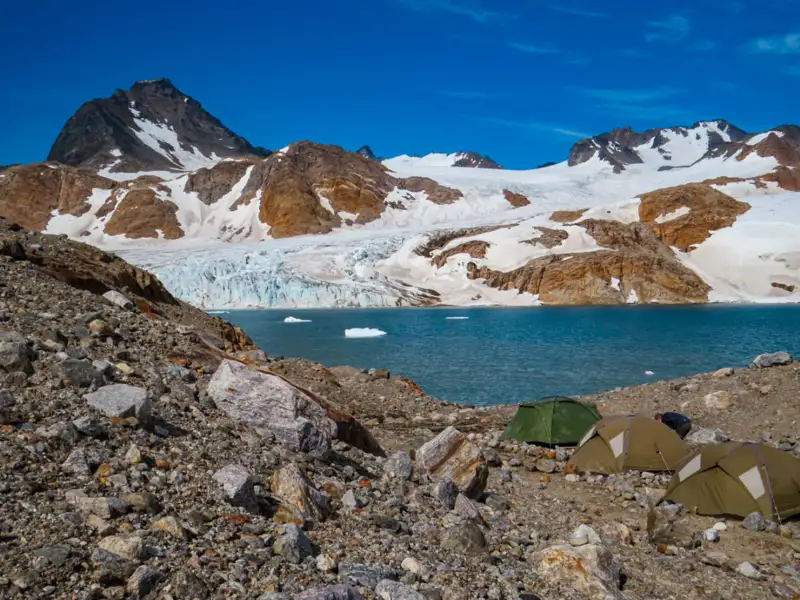 Campingplatz mit Zelten am Ufer eines türkisfarbenen Gletschersees, umgeben von einem Gletscher und schneebedeckten Bergen.