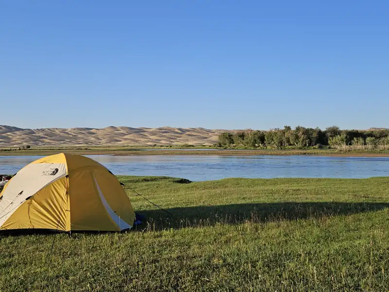 Campingplatz am Fluss mit Zelt und Hügeln im Hintergrund.
