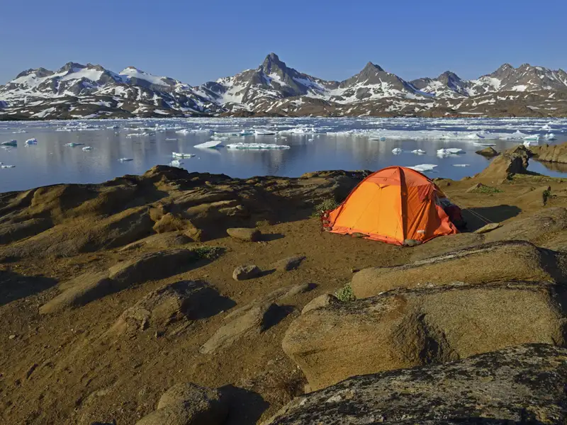 Campingplatz mit Zelt am Ufer eines Fjords in arktischer Umgebung, mit Blick auf schneebedeckte Berge und Eisberge.