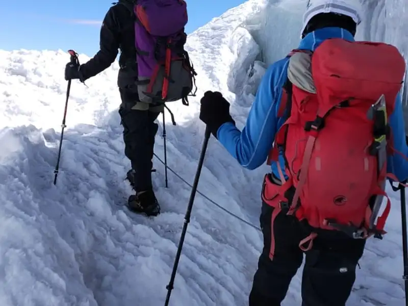 Zwei Personen beim Bergsteigen im Schnee, ausgerüstet mit Rucksäcken und Wanderstöcken. Ein Fixseil dient zur Sicherung.