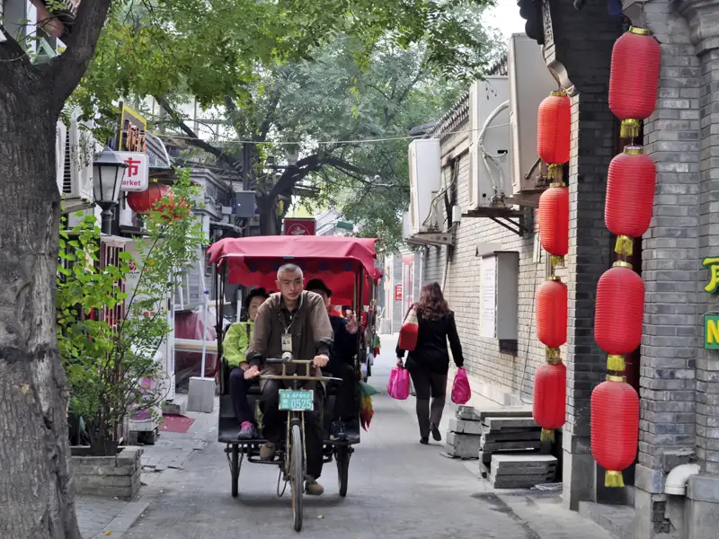 Rikscha-Fahrer transportiert Fahrgäste durch eine enge Gasse, vorbei an traditionellen Gebäuden und roten Laternen.