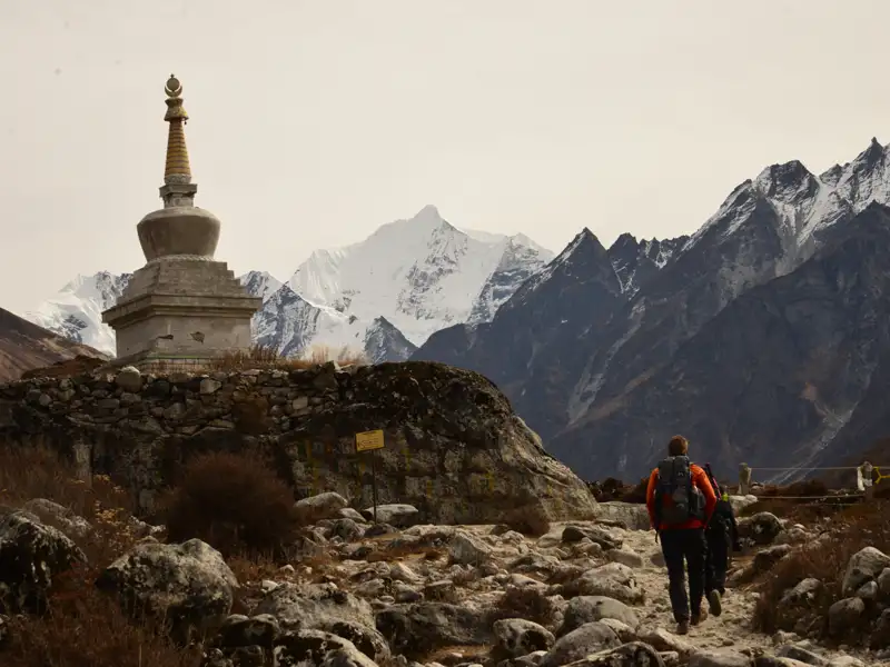 Wanderer auf einem Bergpfad mit einem Stupa und schneebedeckten Bergen im Hintergrund.
