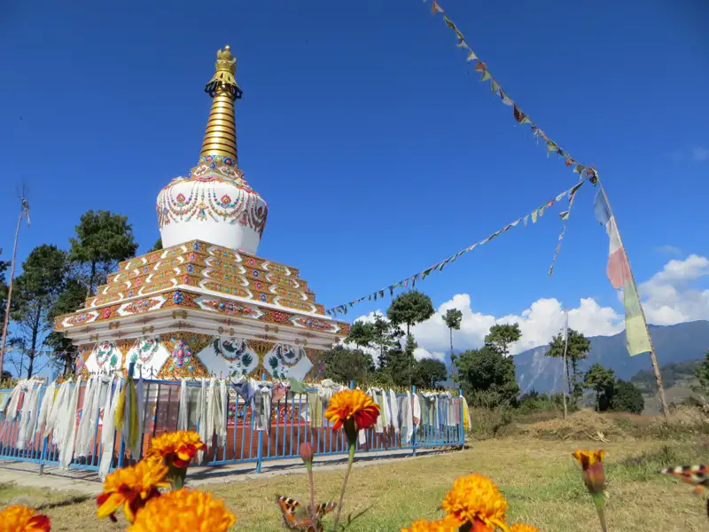 Weißer Stupa mit goldenen Verzierungen und bunten Gebetsfahnen, umgeben von einem blauen Zaun und orangen Blumen. Im Hintergrund Bäume und Berge.
