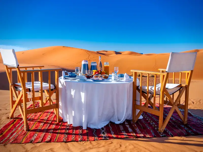 Frühstück in der Wüste, serviert auf einem Tisch mit weißer Tischdecke und zwei Stühlen auf einem Teppich. Im Hintergrund sind Sanddünen zu sehen.