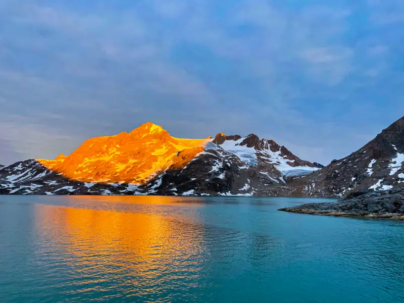 Schneebedeckte Berge und Gletscher im goldenen Abendlicht, die sich im türkisfarbenen Wasser spiegeln.