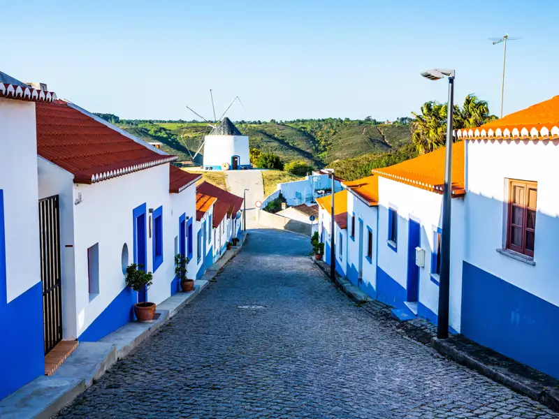 Weiß-blaue Häuser säumen eine gepflasterte Straße, die zu einer Windmühle auf einem Hügel führt.