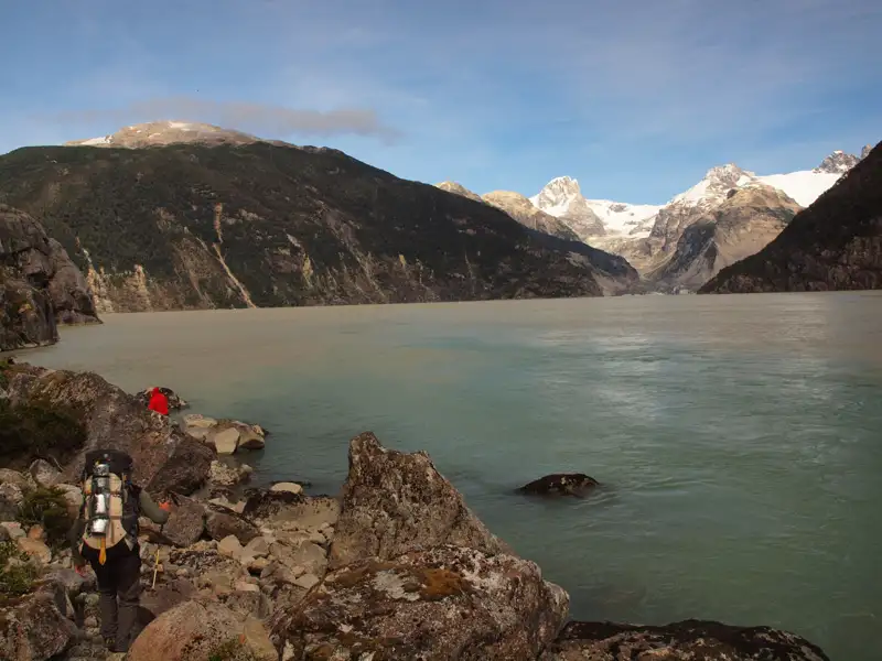 Wanderer am Ufer eines Gletschersees mit Bergen und Gletschern im Hintergrund.