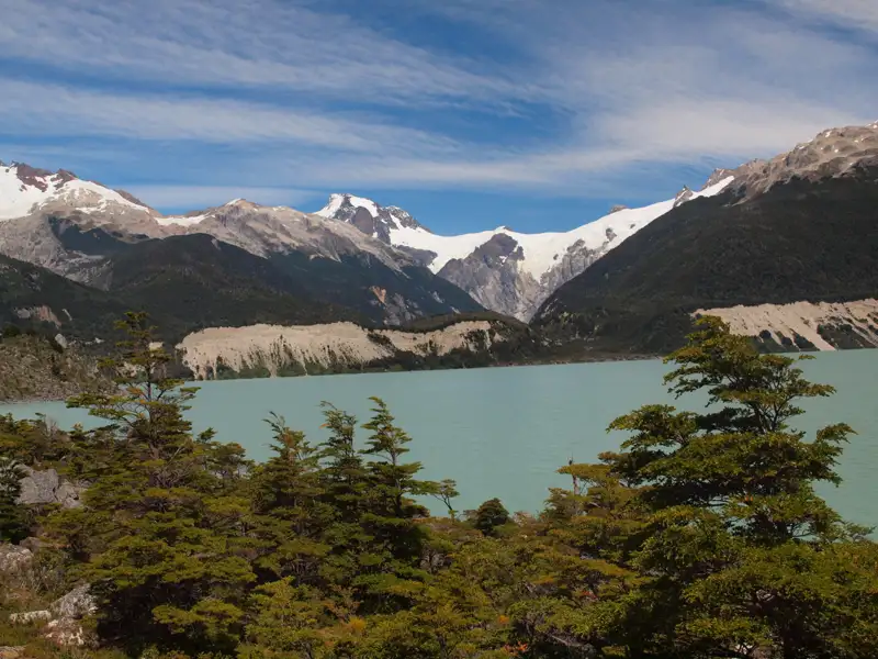 Panorama eines türkisfarbenen Sees mit umgebender Vegetation und schneebedeckten Bergen im Hintergrund.