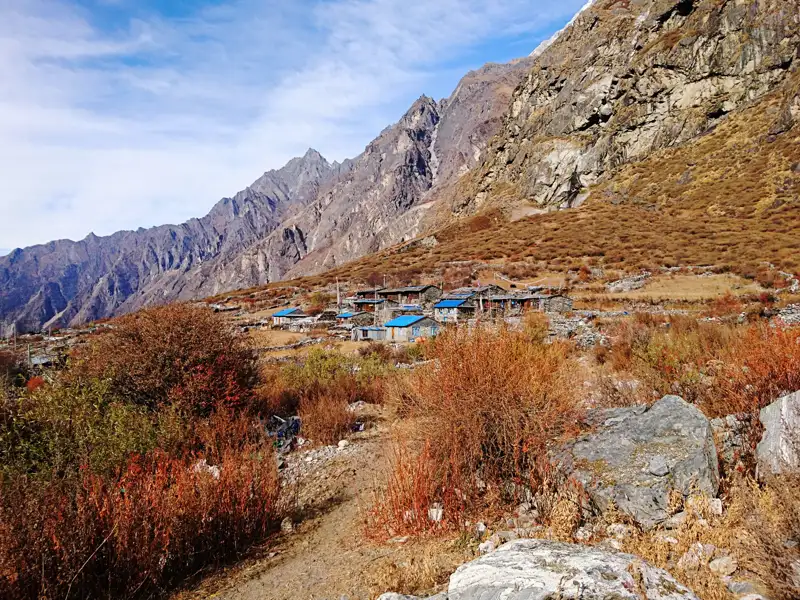 Kleines Bergdorf mit blauen Dächern, umgeben von herbstlicher Vegetation und felsiger Landschaft.