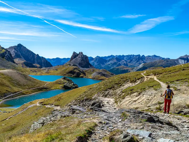 Wanderer auf einem Bergpfad inmitten einer Gebirgslandschaft mit türkisblauen Seen.