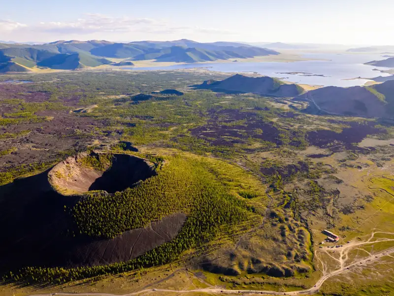 Luftbild eines Vulkankraters mit umliegender Vegetation, einem See im Hintergrund und Anzeichen von vulkanischer Aktivität in der Landschaft.