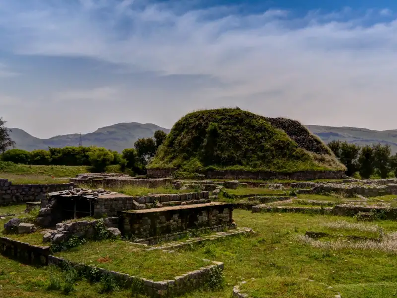 Panoramablick auf die historischen Ruinen mit einem begrasten Hügel und Bergen im Hintergrund.