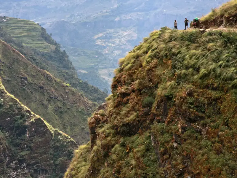 Zwei Personen auf einem Berggrat mit Blick auf ein tiefes Tal und terrassierte Felder.