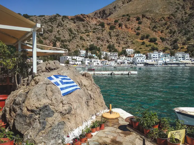 Griechische Flagge auf einem Felsen, mit Blick auf den Hafen und die weißen Häuser im Hintergrund.