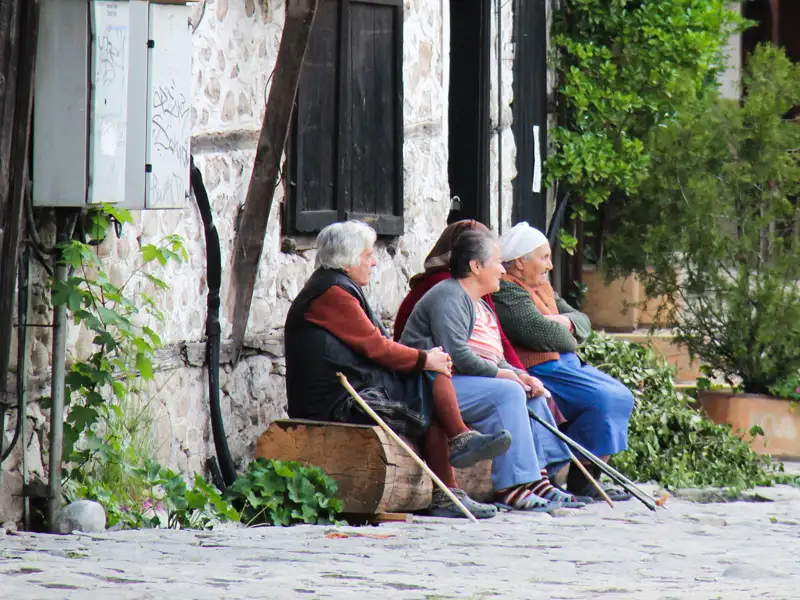 Drei ältere Frauen ruhen sich auf einer Bank vor einem Steinhaus aus.
