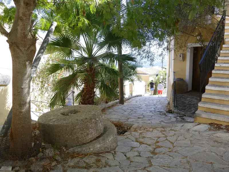 Gepflasterter Weg in einem griechischen Dorf mit altem Mühlstein und Palmen.
