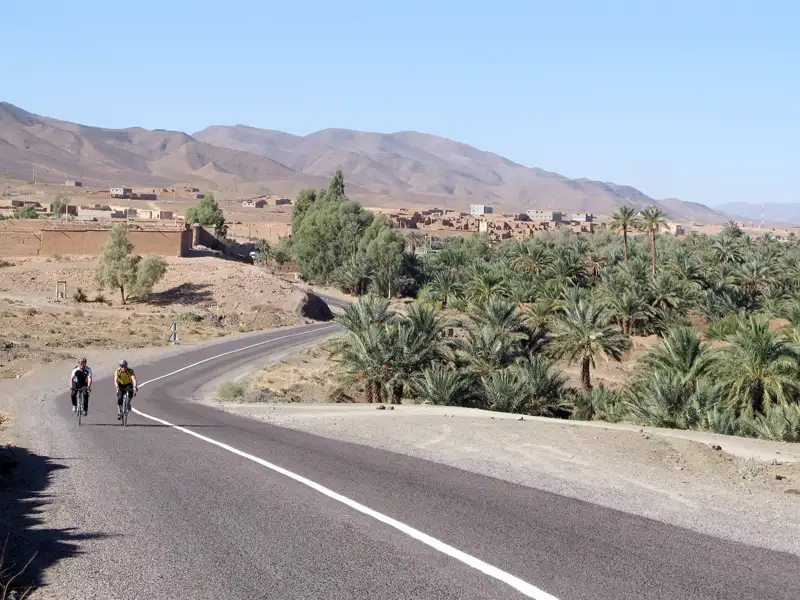 Radfahrer auf einer Straße in der Nähe einer Oase und eines Dorfes in einer bergigen Wüstenlandschaft.