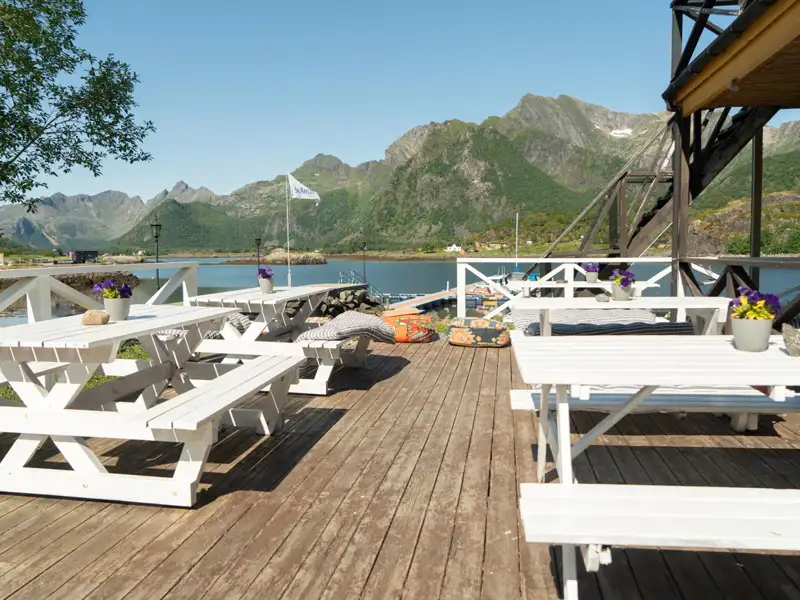 Picknicktische auf einer Holzterrasse mit Blick auf einen Fjord und Berge im Hintergrund.