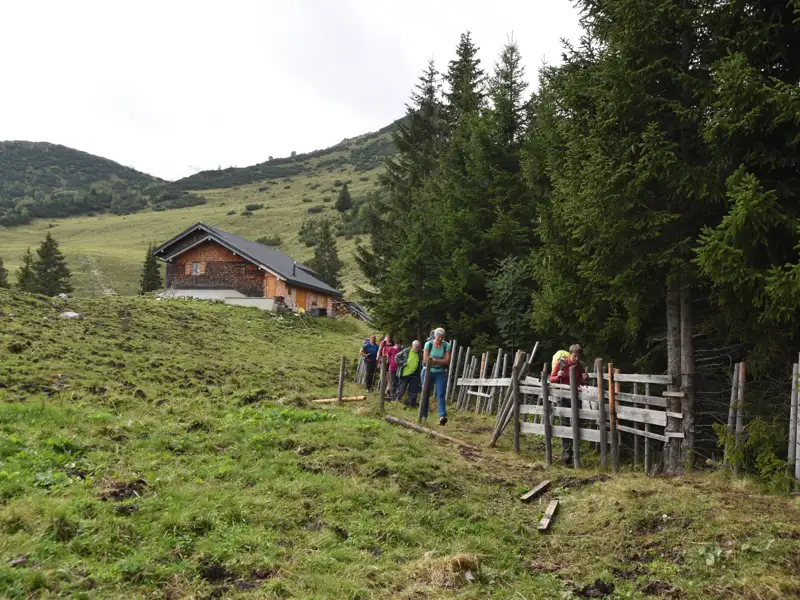 Wanderer auf einem Bergpfad, die einen Weidezaun passieren und sich einer Berghütte nähern.