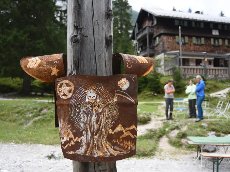 Rostiges Metallschild mit Sensenmann und Bergen, vor einer Berghütte.