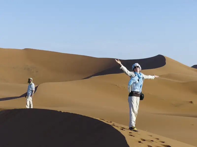 Zwei Reisende in einer sandigen Wüstenlandschaft. Eine Person steht mit ausgebreiteten Armen auf einer Düne.