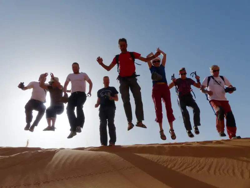 Reisegruppe springt freudig über eine Düne in der Wüste.